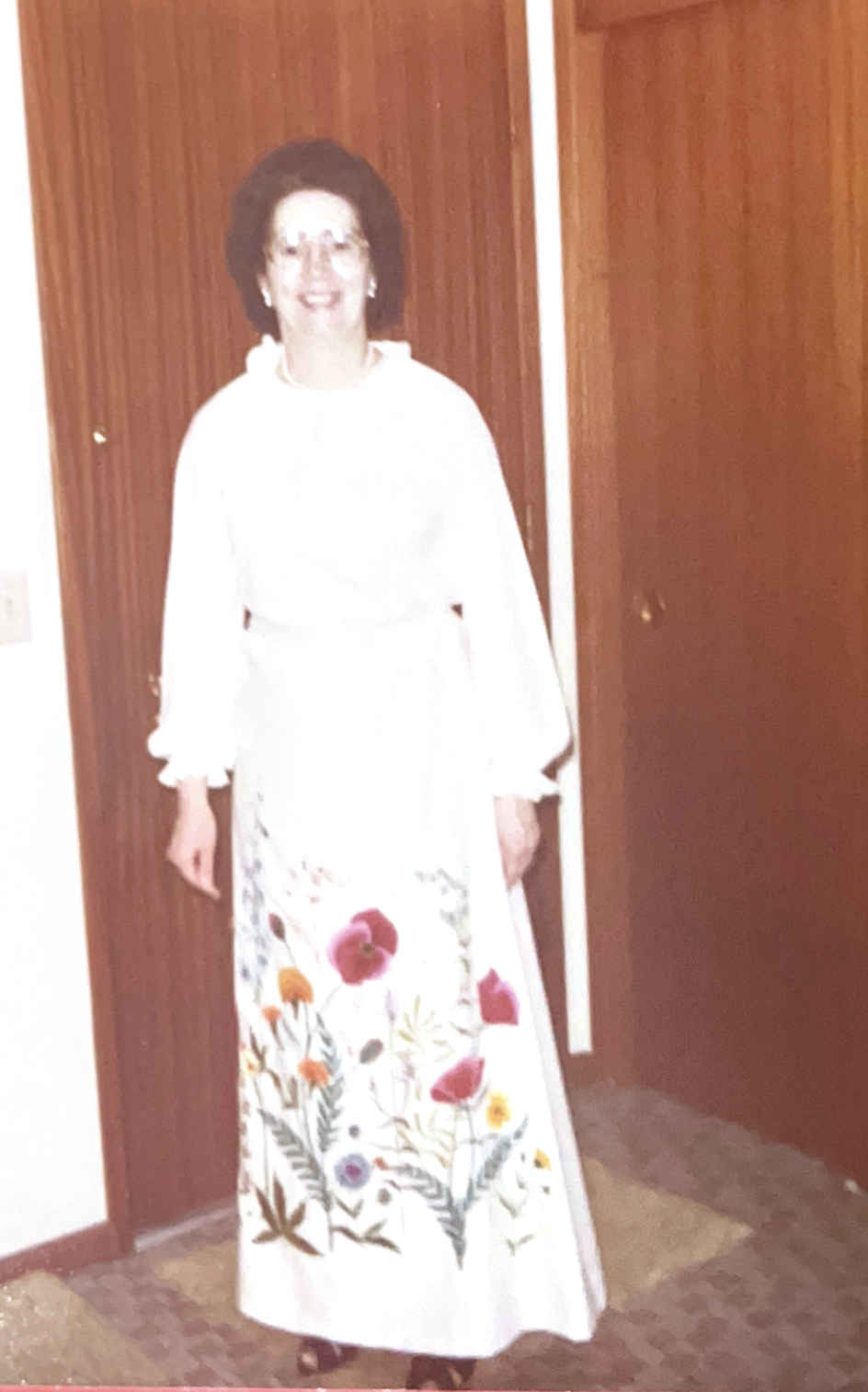 A picture from the 1970s of a woman wearing a white blouse and skirt. The skirt is long and covered with embroidered flowers. Wood doors are in the background.